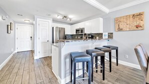 A modern kitchen with white cabinets, stainless steel appliances, and a bar area with stools. - Gulf Views! Ground Unit! Patio! Ciboney 1002! (Miramar Beach)