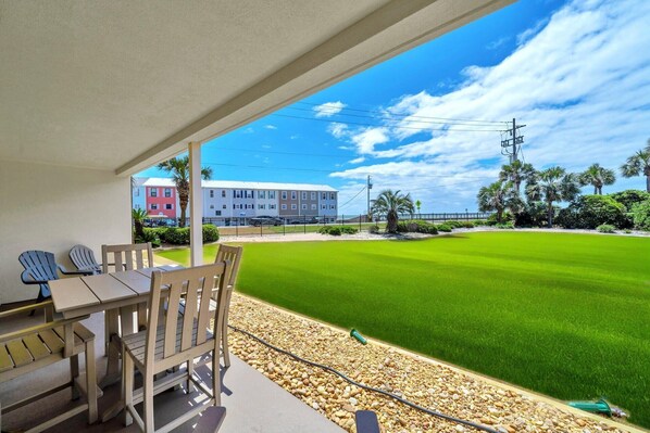 A covered patio with a table and chairs, a well-maintained lawn, and a building with multiple stories in the background.