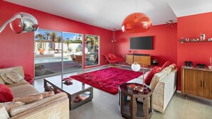 A modern living room with red walls, a glass coffee table, a red rug, and a view of a pool and outdoor furniture. - Designer Wexler Oasis with Saltwater Pool & Spa (Palm Springs)