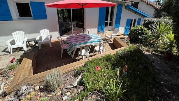 A patio with a table and chairs, a red umbrella, and a wooden deck. - 032 - 3 *** villa in a quiet area close to the beaches and the city center (SOULAC SUR MER)