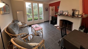 A cozy living room with wicker furniture, a fireplace, and large windows offering a view of greenery. - 032 - 3 *** villa in a quiet area close to the beaches and the city center (SOULAC SUR MER)