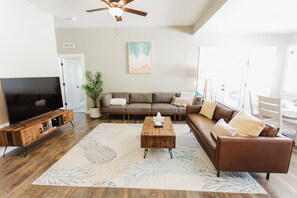 A living room with a large flat-screen TV, a wooden coffee table, a brown leather sofa, and a large abstract rug. - Beautiful Home with HOT TUB, COMMUNITY POOL & GAME ROOM. (Nampa)