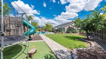 A backyard with a trampoline, a green slide, a small wooden bench, a stone pathway, and a fenced area with trees.
