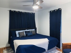Ein Schlafzimmer mit einem Bett, dunklen Vorhängen, einem Deckenventilator und einer weißen Tagesdecke.
