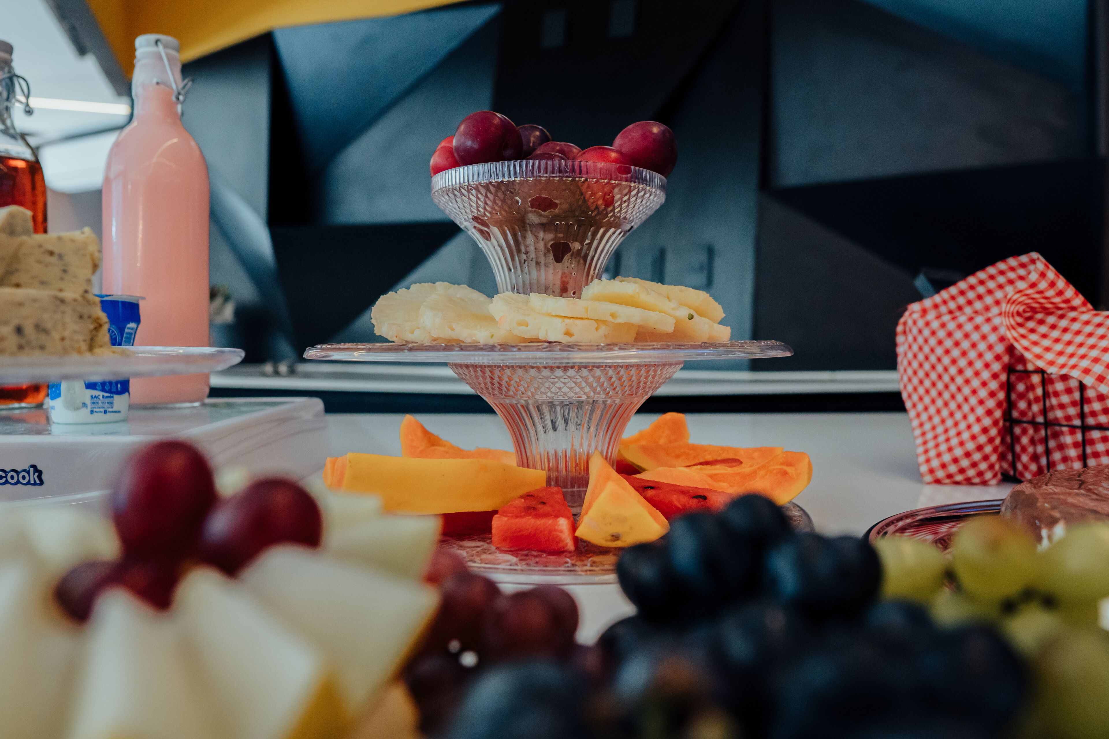 Eine Obst- und Käseplatte mit einer Flasche pinkfarbenem Getränk und einer karierten Serviette auf einem Tisch.