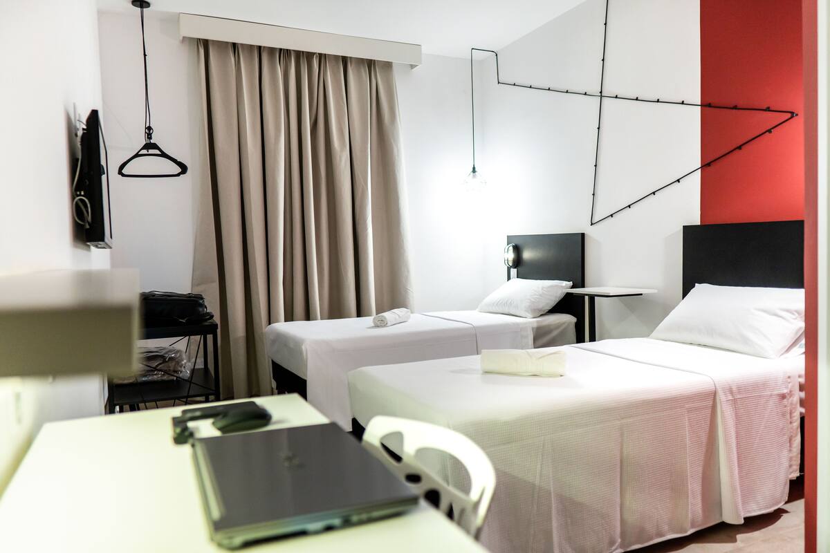 Ein Hotelzimmer mit zwei Betten, einem Schreibtisch mit einem Laptop, einem Sessel, einem Fernseher und einer roten Akzentwand.