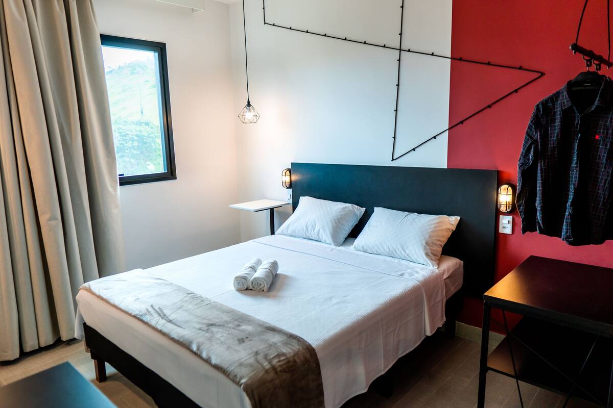 Ein ordentlich gemachtes Bett mit weißer Bettwäsche und einem zusammengerolltem Handtuch, ein Nachttisch mit Lampe und eine Wand in roter Farbe.