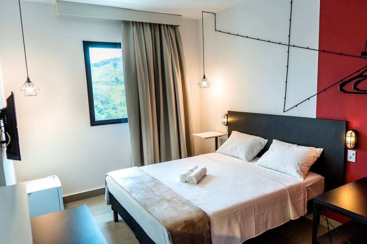 Ein Hotelzimmer mit Bett, Nachttisch, Lampe und Blick auf Grün durch das Fenster.