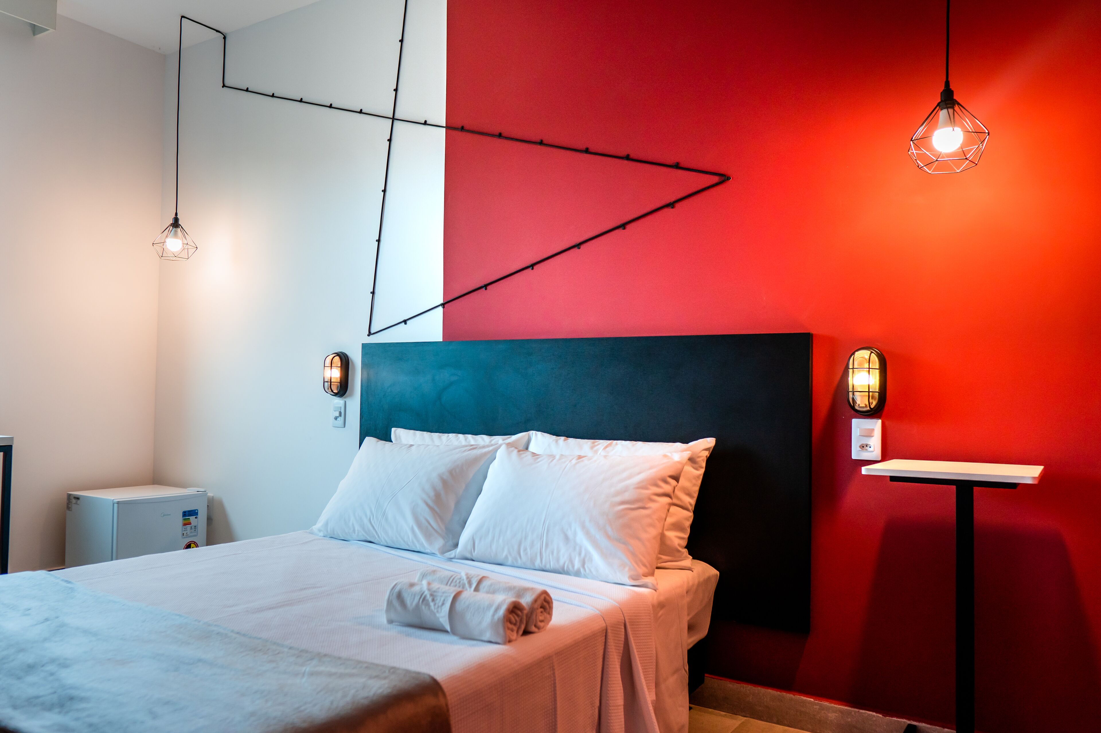 Een moderne hotelkamer met een bed, witte lakens, een rode accentmuur en hanglampen.