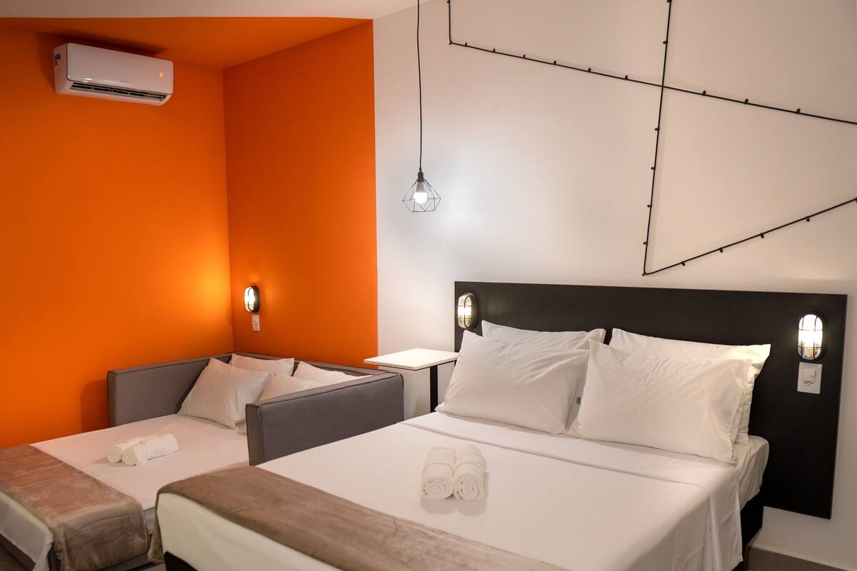 Ein Hotelzimmer mit einem großen Bett, einem kleineren Bett und einem Schlafsofa. An der Wand ist eine Klimaanlage angebracht und eine Pendelleuchte hängt von der Decke.