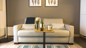 Un salon avec un canapé beige, deux coussins, une table basse en bois sur laquelle se trouvent une bouteille, des verres et une bougie. Deux œuvres d'art encadrées représentant des plumes sont accrochées au mur.