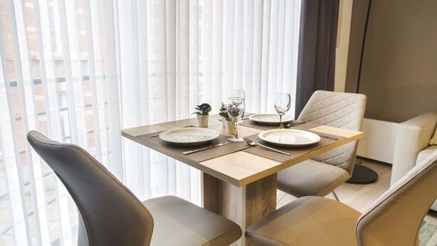 Un coin repas avec une table dressée pour deux personnes, deux chaises, une petite plante en pot et un verre d’eau.
