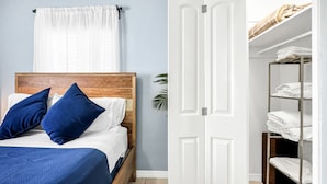 Ein ordentlich bezogenes Bett mit blauer Bettwäsche und Kissen, eine weiße Tür mit Metallgriff und ein Metallregal mit gefalteten Handtüchern.
