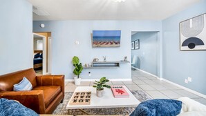 Ein Wohnzimmer mit braunem Ledersofa, einem Couchtisch mit Buch über Palmen und einem Holz-Gesellschaftsspiel, einem Fernseher an der Wand und einem Gemälde mit Strandmotiv.