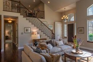 A spacious living room with a staircase, a chandelier, and a view of the outdoors. - Village Greens View (Kalispell)