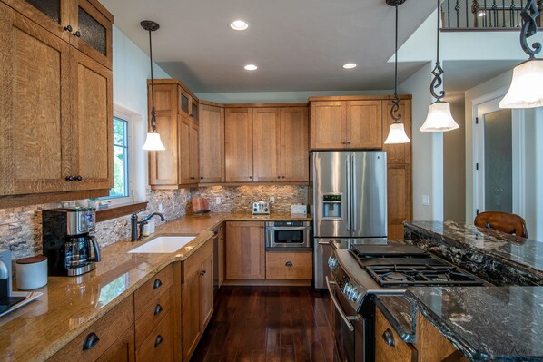 A modern kitchen with wooden cabinets, granite countertops, stainless steel appliances, and pendant lighting. - Village Greens View (Kalispell)