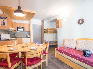 A compact room with a wooden dining table, chairs, a bunk bed, a kitchenette, and a small seating area.