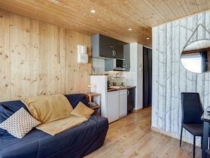 A cozy room with a wooden ceiling, a dark blue sofa with a yellow throw, a small table with a lamp, and a view of a kitchen and dining area through an open door. - Cosy apartment for 2 people with WIFI, TV and balcony (Les Boisses)