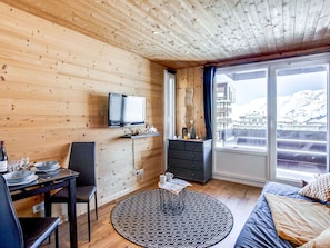 A cozy room with wooden walls, a TV mounted on the wall, a dining table with chairs, a sofa, and a large window offering a view of a snowy landscape. - Lovely apartment for 2 people with WIFI, TV and balcony (Les Boisses)