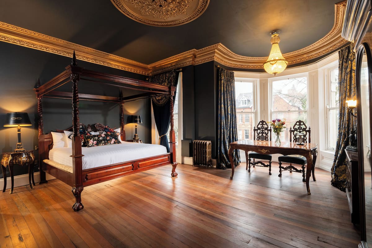 A four-poster bed with a wooden frame, a bedside table with a lamp, and a chair at a desk with a vase of flowers.