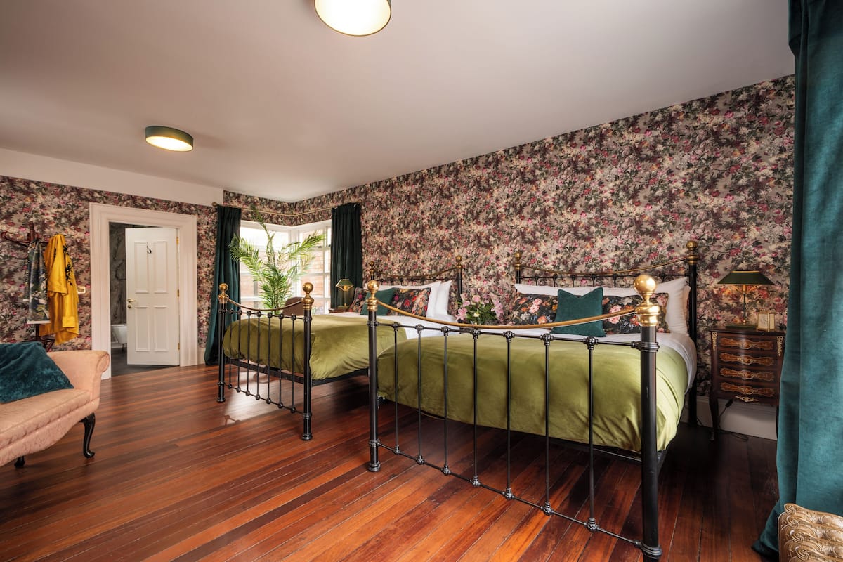 A bedroom with two beds, wooden flooring, floral wallpaper, and a green curtain.
