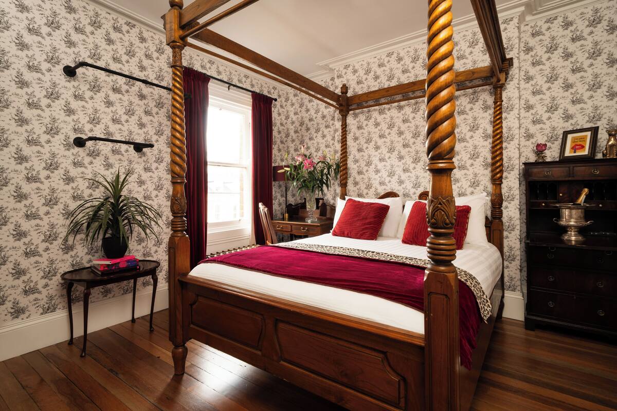 A four-poster bed with a wooden frame and a red bedspread in a room with floral wallpaper and a wooden floor.