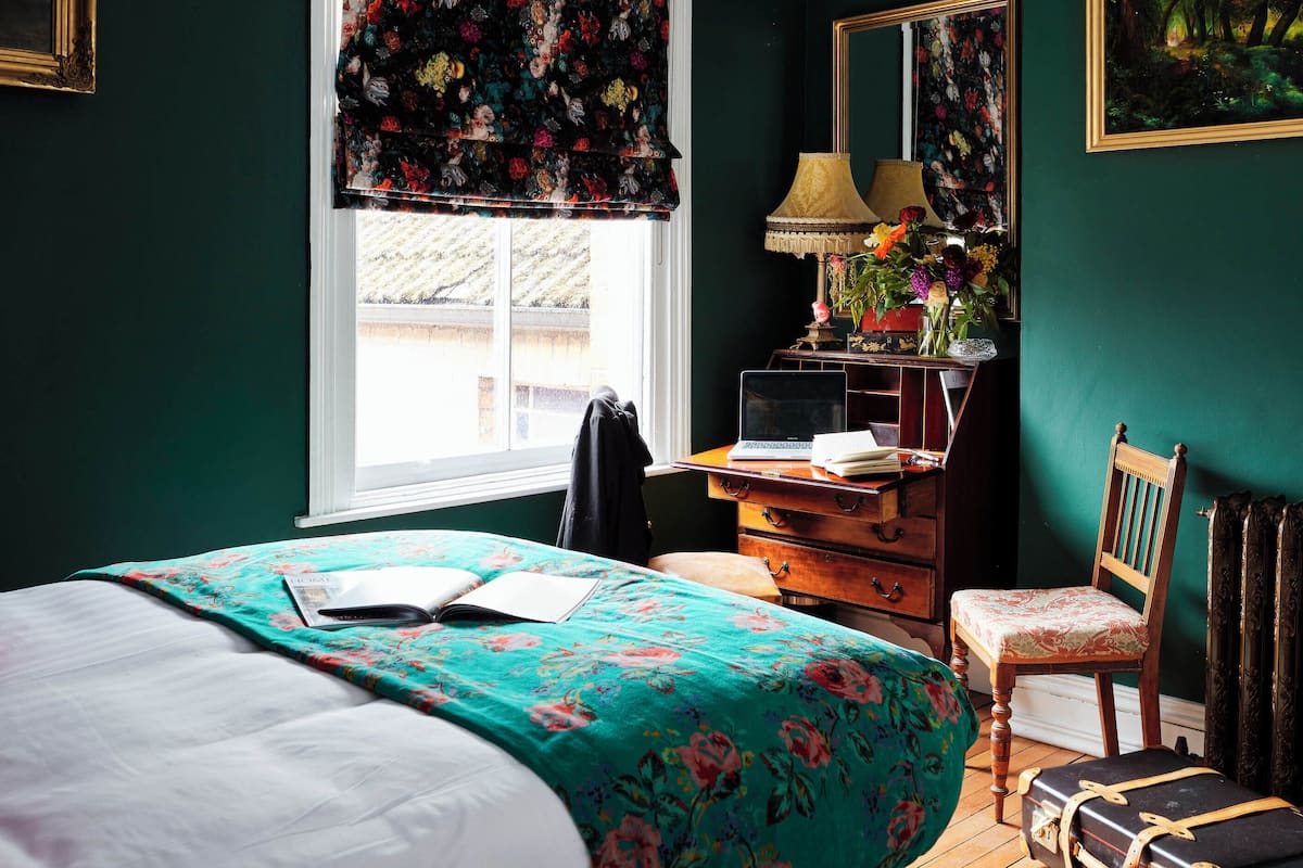 A bedroom with a bed, a desk, a chair, a lamp, and a window with floral blinds.