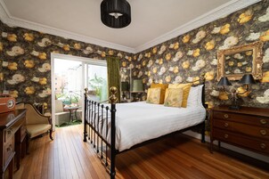 Un dormitorio con cama, suelo de madera, papel tapiz floral, cómoda, silla, mesa y ventana con vistas.