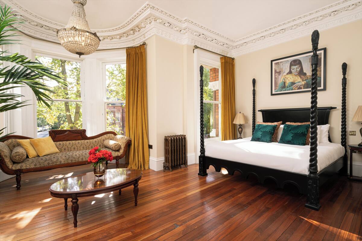 A room with a four-poster bed, a sofa, a wooden coffee table, and a large window with curtains.