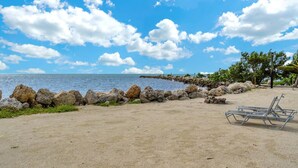 Uma praia com um paredão rochoso, céu azul claro e algumas cadeiras de praia.