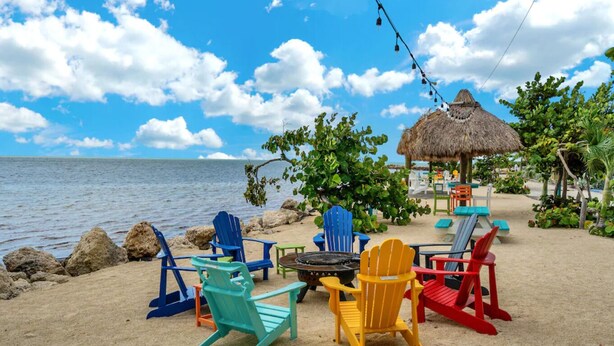 Cenário de praia com cadeiras Adirondack coloridas, uma estrutura com telhado de palha e vista para o oceano.
