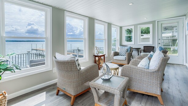 A spacious room with large windows overlooking a body of water, wicker furniture, and a small table. - Petit Retreat: The sweetest place on the Lake! (Seneca Falls)