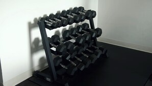 A black dumbbell rack with multiple pairs of dumbbells against a white wall. - Whitewood Inn (Whitewood)