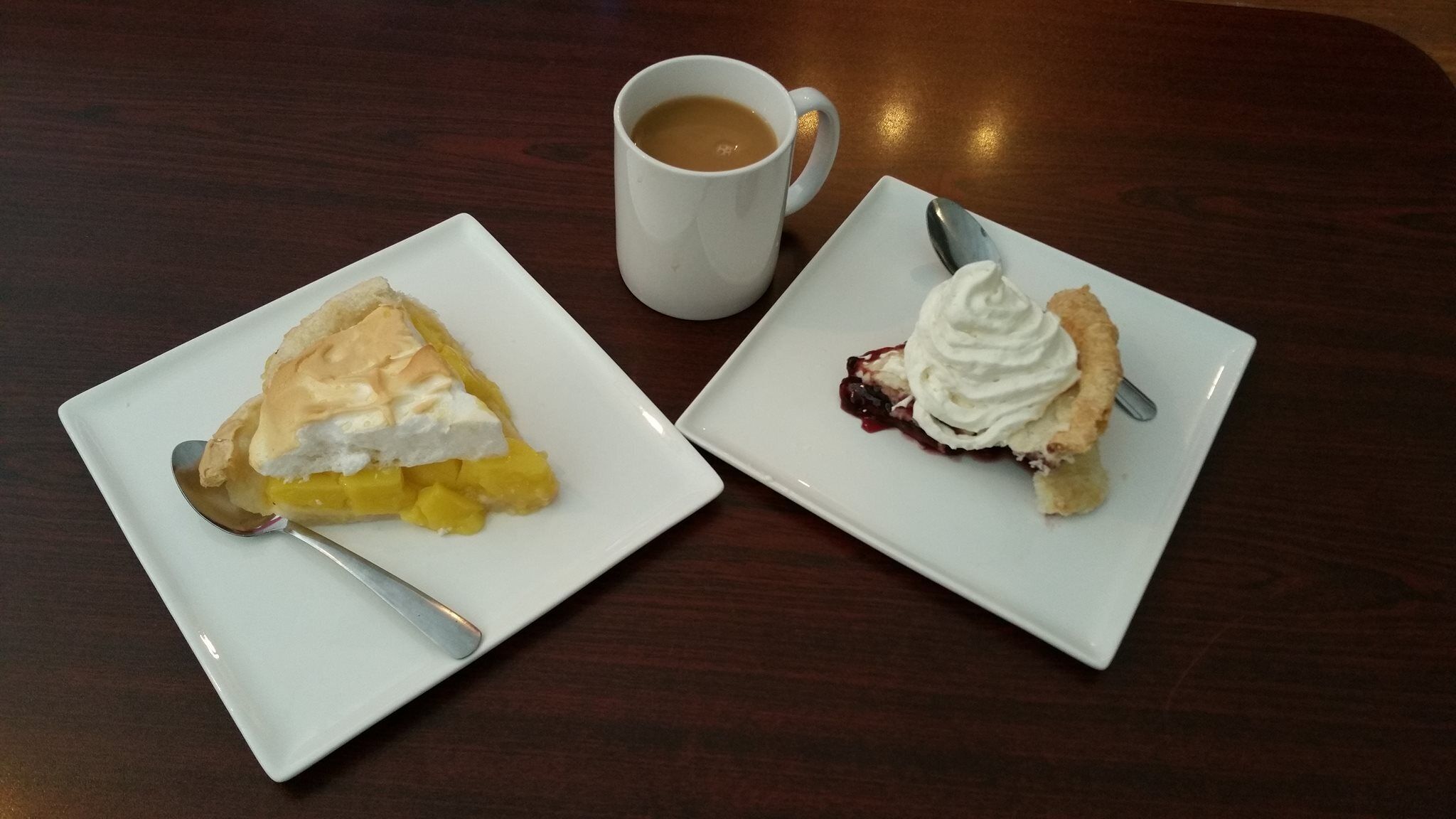 Two square white plates with slices of pie, one with mango and the other with a berry topping, and a white mug with a spoon on a wooden table.