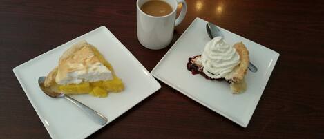 Dos platos blancos cuadrados con porciones de tarta, una con mango y otra con cobertura de frutos rojos, y una taza blanca con una cuchara sobre una mesa de madera.
