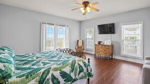 Quarto com cama, ventilador de teto, cômoda, televisão e janelas amplas.