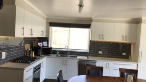 A modern kitchen with white cabinets, a black tiled backsplash, a window with blinds, and a dining area with a wooden table and chairs. - PeliCann Sands Beach House - Loch Sport (Loch Sport)