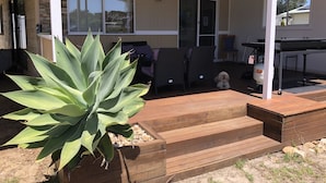 A covered patio area with a wooden deck, outdoor furniture, and a large agave plant. - PeliCann Sands Beach House - Loch Sport (Loch Sport)