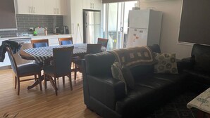 A modern kitchen with white cabinets, a dining table with chairs, a black leather sofa, and a small table with a tablecloth. - PeliCann Sands Beach House - Loch Sport (Loch Sport)