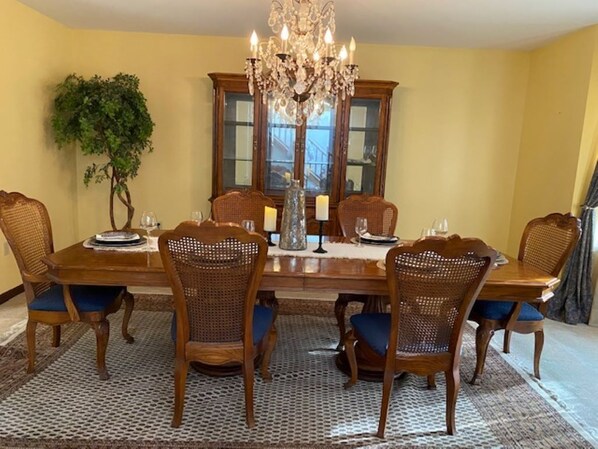 A dining room with a wooden table, six wicker chairs, a chandelier, and a large window with a view.