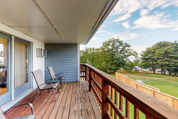 Relax on the deck with a view of lush green surroundings, surrounded by a railing and adjacent to a building with a window and air conditioning unit.