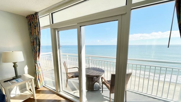 Een kamer met een grote glazen schuifdeur die uitkomt op een balkon met uitzicht op de oceaan. Op het balkon staan een klein tafeltje en twee stoelen.