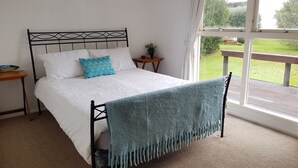 Una cama con cubrecama blanco y manta azul, una mesita de noche con una planta, y vista a un área verde por la ventana.