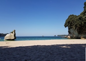 Une plage à l’eau d’un bleu cristallin, une grande formation rocheuse et une falaise couverte d’une végétation luxuriante.