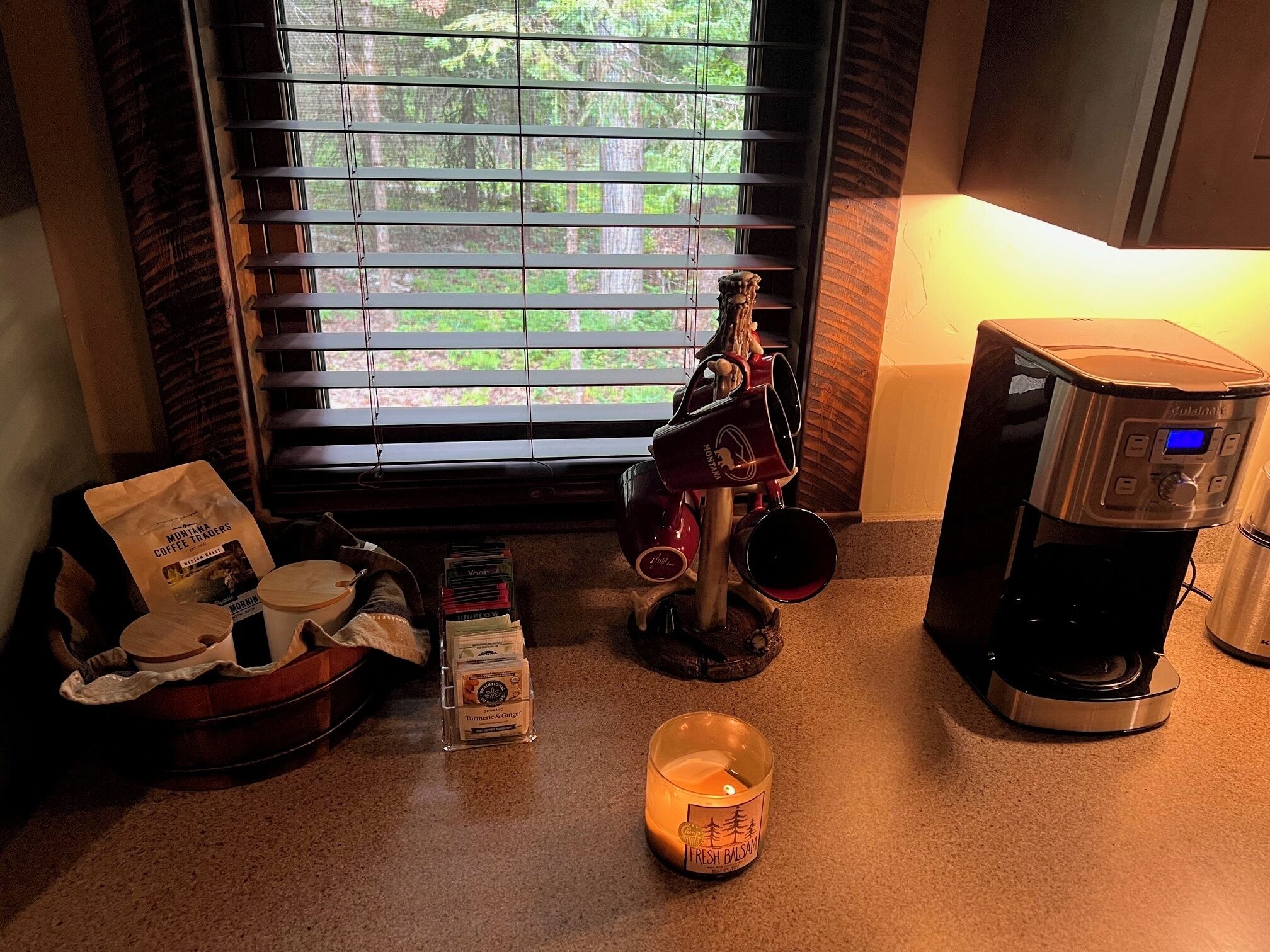 A kitchen area with a coffee maker, a coffee grinder, a candle, and a window with blinds.