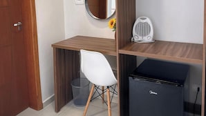 A compact room with a wooden desk, a white chair, a small black refrigerator, a mirror, and a wall-mounted heater. - Pousada Villa Vantino (Penha)