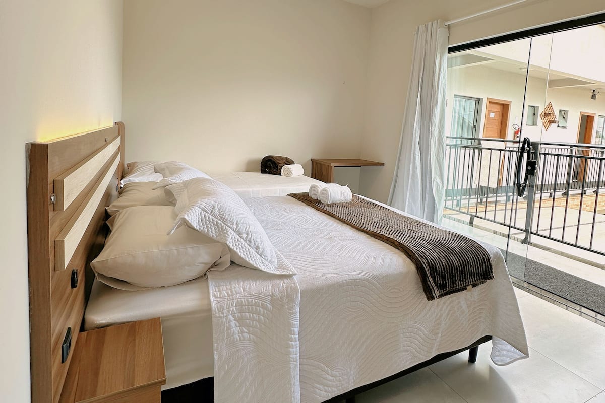 A modern bedroom with a wooden headboard, a large bed with white bedding, a bedside table, and a view of the outside balcony through a glass door.