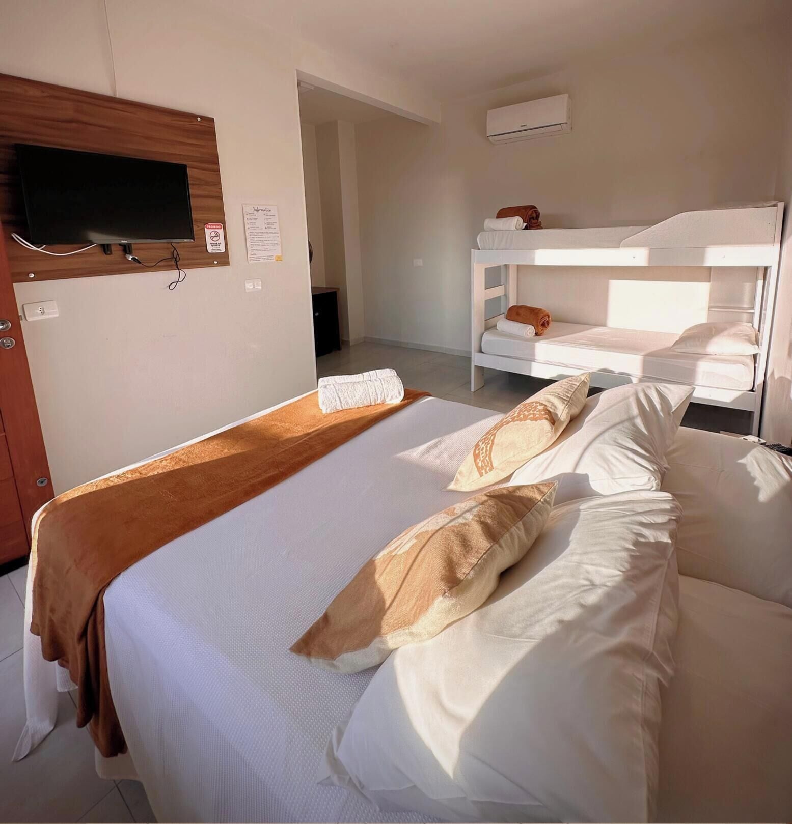 Habitación de hotel con una cama individual, un televisor montado en un panel de madera, una cama litera al fondo y un equipo de aire acondicionado en la pared.