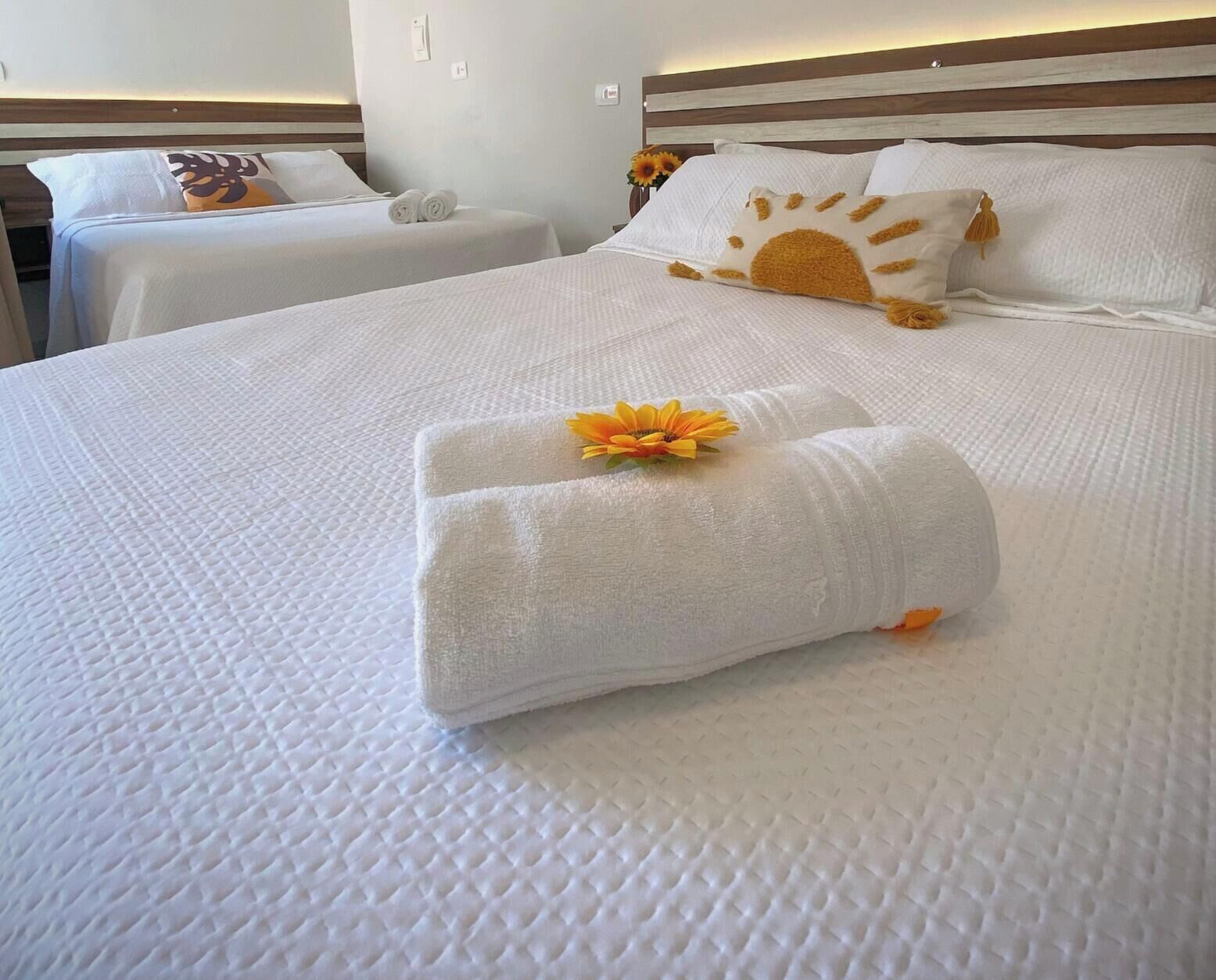 A hotel room with two beds, each with white linens and a sun-shaped pillow. There are towels on the beds and a flower on one of the towels.