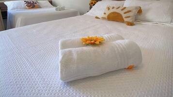 A hotel room with two beds, each with white linens and a sun-shaped pillow. There are towels on the beds and a flower on one of the towels.
